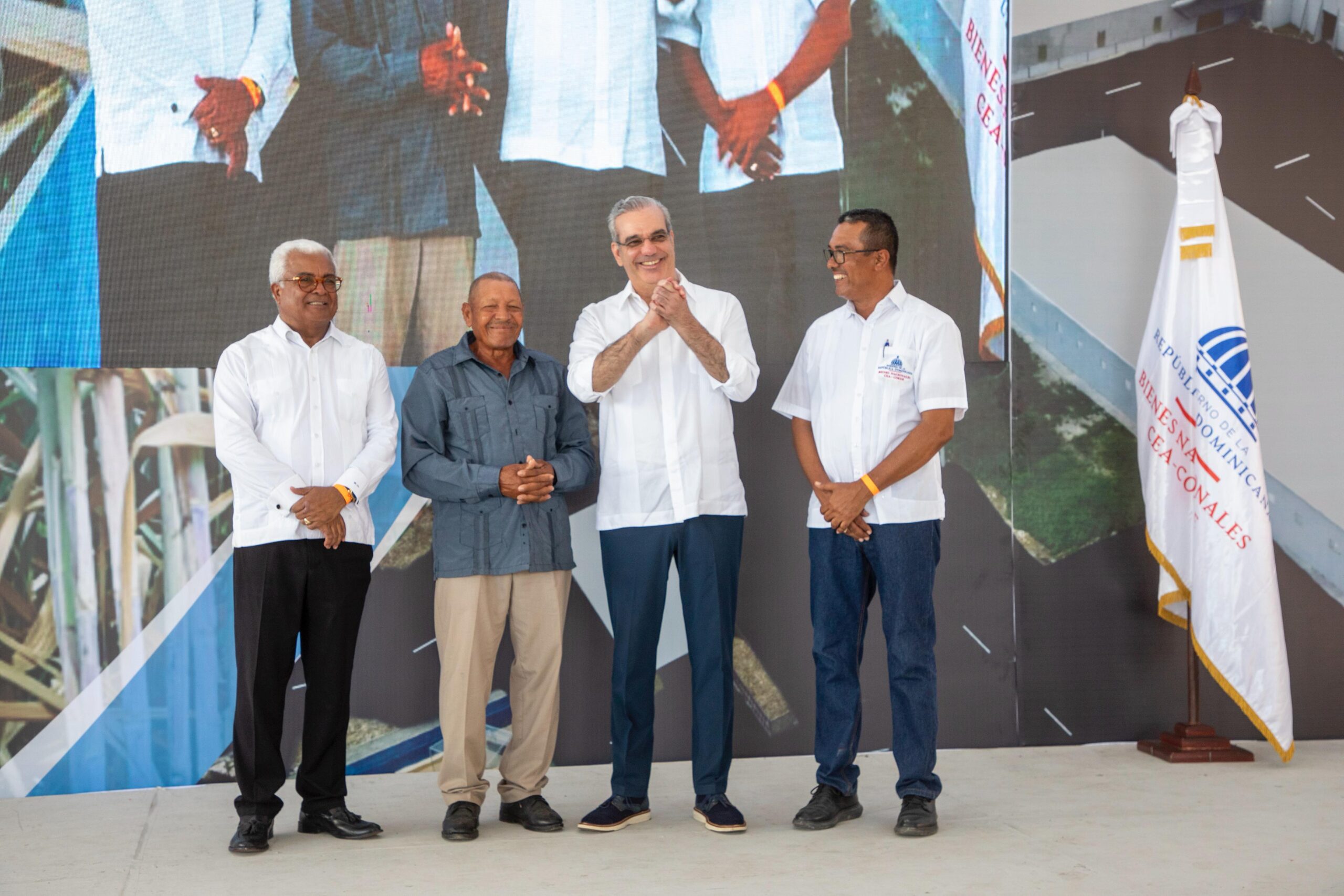 Hombres sonrientes en evento oficial con bandera dominicana.