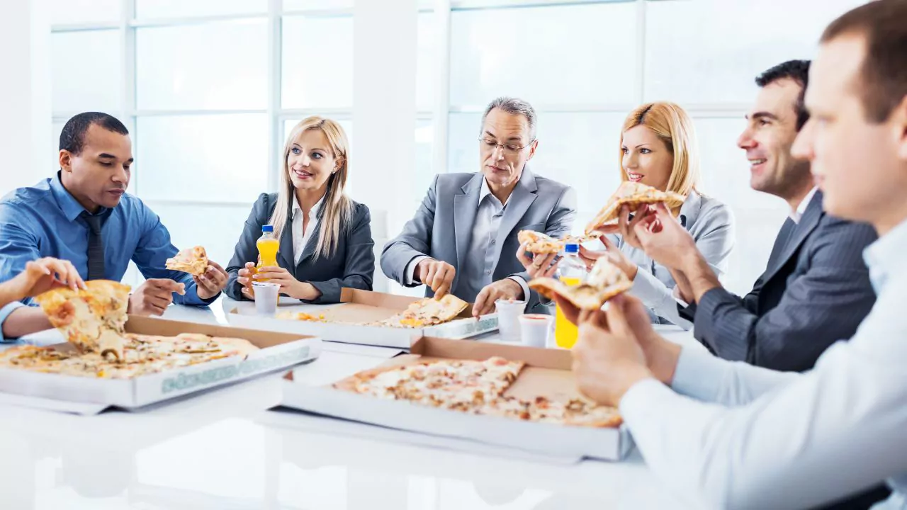 Grupo de empleados almuerza pizza en una oficina, contexto de recomendaciones sobre horarios de comida y energía.